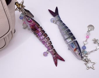 May include: Two fish-shaped keyrings with silver fixings and decorative charms. One is purple and pink, the other is silver and blue. The keyrings are attached to a white handbag.