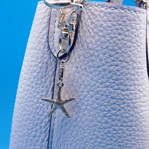 May include: A light blue leather handbag with a silver starfish charm. The bag has a textured surface and silver hardware. A small, matching pouch is attached to the bag. The background is a solid blue.