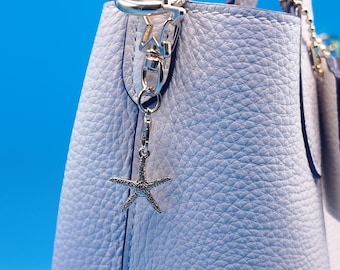 May include: A pale blue leather handbag with a silver starfish charm. The bag has a textured surface and silver hardware. A small, matching pouch is attached. The background is a solid blue.