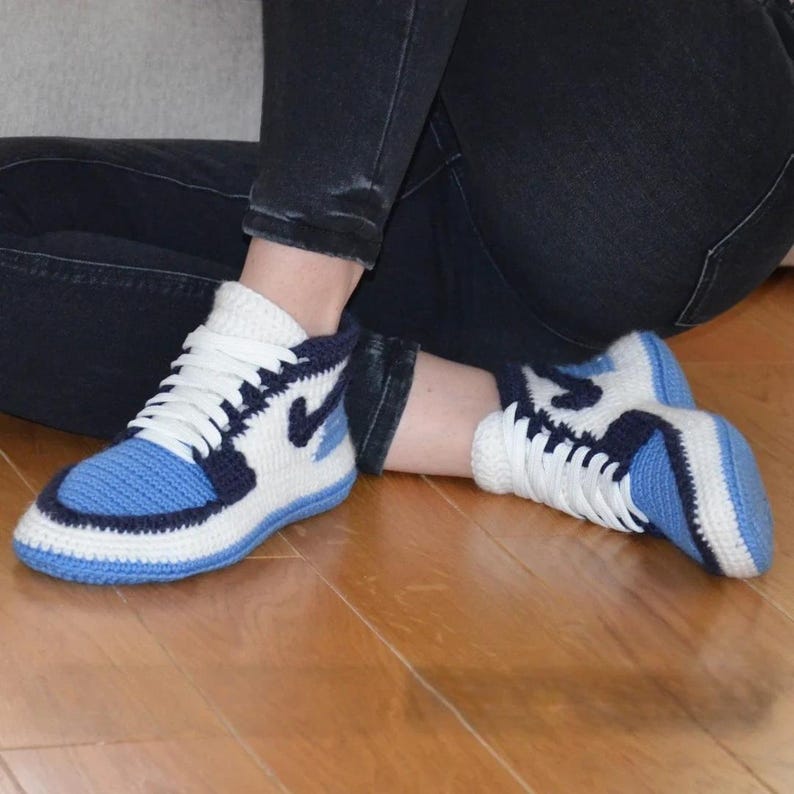 May include: Crocheted sneakers in a high-top style, featuring a blue, white, and navy color scheme. The design mimics a popular athletic shoe, with white laces and a blue swoosh. The shoes are made of yarn.