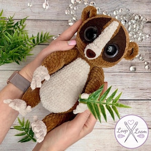 May include: A handmade brown and white crochet lemur plush toy with large black eyes and a white stripe on its face. The plush is holding a green leaf and is being held in two hands. The Loop & Loom logo is in the bottom right corner.