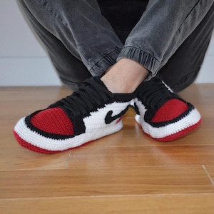 May include: Crocheted sneakers in a classic red, black, and white color scheme. The handmade shoes feature a red toe box, black laces, and a white body with a black swoosh. The sneakers are a cozy, unique take on a popular style.