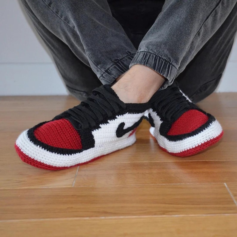 May include: Crocheted sneakers in a classic red, black, and white color scheme. The handmade shoes feature a red toe box, black laces, and a white body with a black swoosh. The sneakers are a cozy, unique take on a popular style.