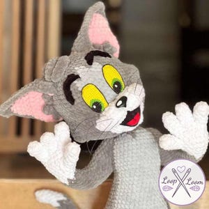 Peut inclure: Une peluche grise en forme de chat avec des oreilles roses, des yeux jaunes et une bouche rouge. Le chat a des pattes blanches et une expression souriante. Le jouet est fait à la main et possède un logo en bas à droite.