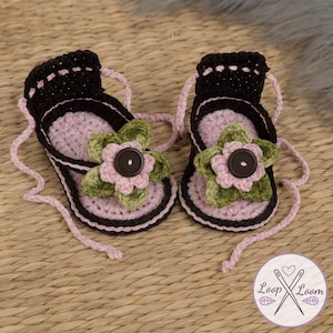 May include: A pair of handmade crochet baby sandals. The sandals are black, pink, and brown with a flower and button detail. The sandals have pink straps and are displayed on a woven surface.
