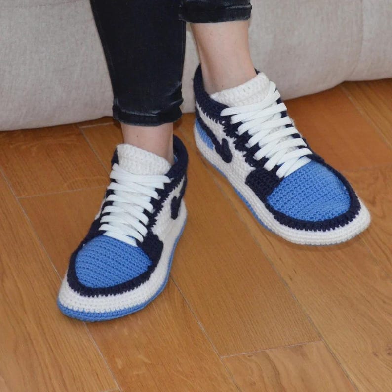 May include: A pair of handmade crochet slippers designed to resemble high-top sneakers. The slippers feature a blue toe box, navy blue accents, white laces, and a white swoosh logo. The slippers are on a wooden floor.