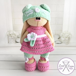 Puede incluir: Muñeca de conejo de ganchillo hecha a mano con un vestido rosa, pantalones verdes y zapatos rosas. El conejo lleva un gorro verde menta con detalles blancos. La muñeca está delante de una pared de ladrillos blancos.