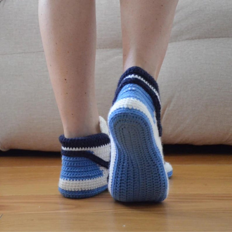May include: Crocheted blue and white slippers. The slippers have a dark blue cuff and are made with a combination of light and dark blue yarn, with white stripes. The slippers are on a wooden floor.