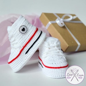 Może przedstawiać: Para białych szydełkowych bucików dla niemowląt z czerwonymi i czarnymi paskami. Jeden but ma czarny guzik w kształcie serca. W tle brązowe pudełko prezentowe z białą wstążką. Widoczne logo Loop & Loom.
