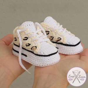 May include: A pair of handmade crochet baby sneakers. The shoes are white with cream-colored accents and black detailing. The sneakers have white laces and a black stripe around the sole. The shoes are small, likely for a newborn.