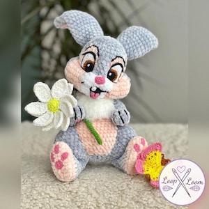 Op de afbeelding: Een handgemaakte pluchen konijn in grijs, roze en wit, met een witte madelief met een geel centrum. Het konijn heeft grote bruine ogen en een roze neus. Een gele en roze vlinder bevindt zich in de buurt van zijn voet. Een zacht speelgoed.