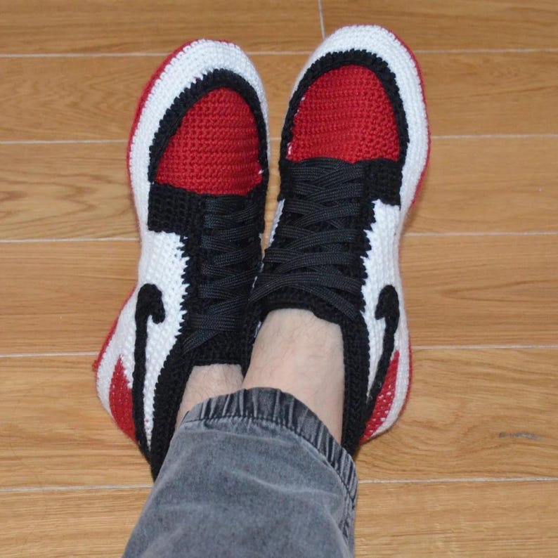 May include: Handmade crochet sneakers in a classic red, black, and white color scheme. The sneakers feature a high-top design with black laces and a black swoosh on the side. The red toe and tongue contrast with the white and black details.