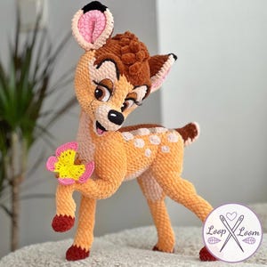 Puede incluir: Figura de ciervo de ganchillo en tonos naranja, marrón y blanco, sosteniendo una flor amarilla y rosa. El ciervo tiene grandes ojos marrones, una nariz negra y orejas internas rosas. La figura está sobre una superficie clara.