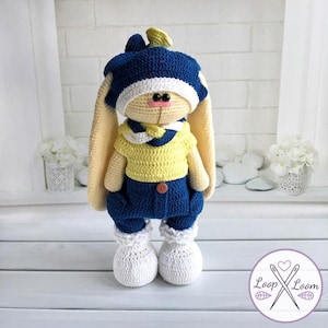 May include: A handmade crochet bunny doll wearing a blue hat, yellow shirt, and blue pants. The bunny has long, beige ears, a pink nose, and white shoes. The doll is standing on a white surface.