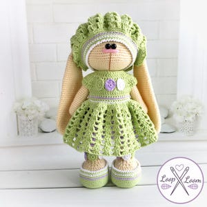 May include: A handmade crochet bunny doll wearing a green dress, matching hat, and shoes. The doll has long, beige ears, black eyes, and a pink nose. The dress features a purple accent and a white flower. The doll is approximately 12 inches tall.