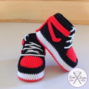 May include: A pair of handmade crochet baby sneakers in a classic high-top design. The shoes feature a black base with red accents on the toe, side, and ankle, and white laces and soles. The sneakers are a charming, handcrafted item.