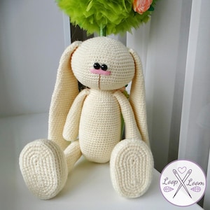 Puede incluir: Un conejo de ganchillo hecho a mano, de color crema, con orejas largas y caídas y una nariz rosa. El conejo tiene ojos de botón negros y está sentado con las piernas extendidas. Es un juguete blando.