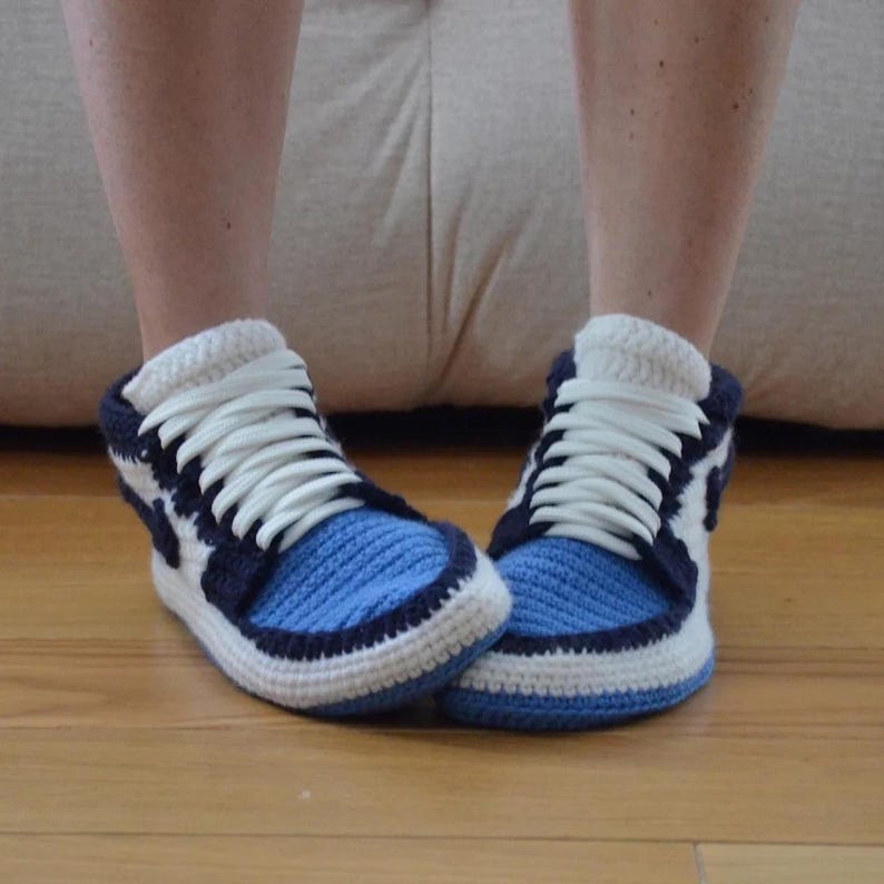 May include: A pair of handmade crochet sneaker slippers. The slippers are blue, white, and navy blue, with white laces. The slippers are on a wooden floor.