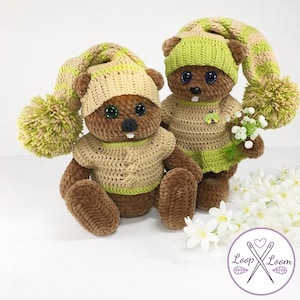 May include: Two handmade crochet teddy bears. Each bear has a brown body, a tan shirt, and a green and tan striped hat with a pom-pom. One bear holds a small bouquet of white flowers. The bears have large, expressive eyes. The outfits are all hand-stitched.