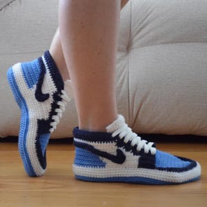 May include: Crocheted blue, white, and navy blue high-top sneaker slippers. The slippers feature a classic sneaker design with a white sole, blue accents, and a black swoosh logo. The slippers are made of soft yarn and have white laces.