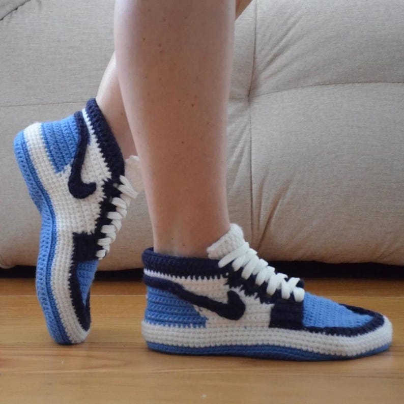 May include: Crocheted blue, white, and navy blue high-top sneaker slippers. The slippers feature a classic sneaker design with a white sole, blue accents, and a black swoosh logo. The slippers are made of soft yarn and have white laces.