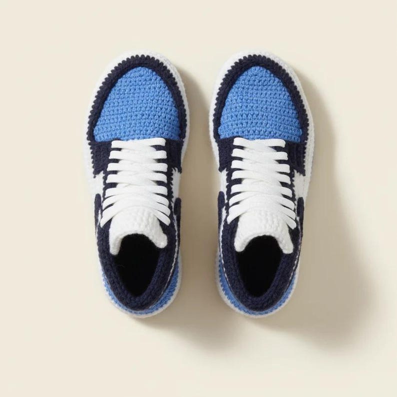 May include: A pair of handmade crochet sneakers in a top-down view. The sneakers feature a blue toe and tongue, white sides, and navy blue accents. White laces are laced up. The shoes are set against a cream-colored background.