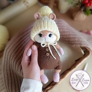 May include: A crocheted animal doll wearing a cream-colored knit hat with a pom-pom and a brown knit romper. The romper features three small wooden buttons down the front and a white ribbon tie at the neck. The doll has light pink paws and feet.