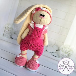 May include: A handmade crochet bunny doll with long ears, wearing a pink outfit and shoes. The bunny has a cream-colored body, a pink nose, and black eyes. The doll is adorned with a pink headband and white flower accents, perfect for a child's toy or nursery decor.