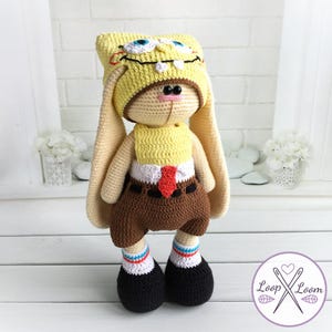 Op de afbeelding: Een handgemaakt gehaakt pluche speelgoed in de vorm van een konijn verkleed als SpongeBob SquarePants. Het speelgoed heeft een gele hoed met geborduurde ogen en tanden, een gele top, bruine shorts en zwarte schoenen met gestreepte sokken.