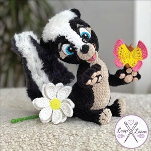May include: A handmade crochet skunk plushie with a white stripe down its back, holding a pink and yellow butterfly. A white daisy with a yellow center and green stem is on the left. The skunk has blue eyes and is smiling.
