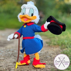 Könnte beinhalten: Gehäkelte Figur einer Zeichentrickente mit blauem Mantel mit roten Verzierungen, rotem Gürtel und roten Stiefeln. Die Ente hat einen weißen Kopf mit weißem Hut, Brille und gelbem Schnabel. Sie hält einen Stock und einen schwarzen Hut.