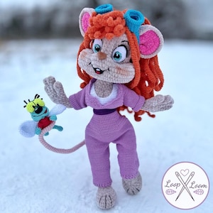 May include: A crocheted doll of a mouse character with long orange hair, blue goggles, and a purple jumpsuit. The mouse has large ears with pink centers and is holding a small, green, winged insect creature with a red body and yellow head.