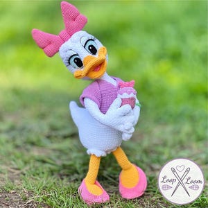 Puede incluir: Muñeca de ganchillo de Daisy Duck hecha a mano, con cuerpo blanco, lazo rosa y camisa morada. La muñeca sostiene una magdalena y lleva zapatos rosas y amarillos. La muñeca está sobre hierba verde.