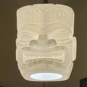 Puede incluir: Una lámpara colgante blanca con forma de máscara Tiki. La luz tiene un diseño tallado con una cara detallada, que incluye ojos, nariz y boca. La luz cuelga de una cuerda marrón.