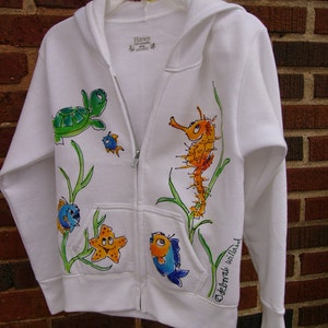May include: White zippered hoodie with a hand-painted design of sea creatures, including a turtle, starfish, seahorse, and fish. The text "Deborah Williams" is written on the right sleeve.