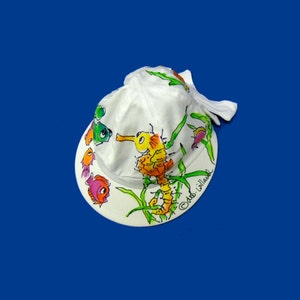May include: White baseball cap with a hand-painted design of a seahorse and fish. The cap has a curved peak and a fabric strap. The artwork includes green seaweed and the artist's signature. The background is a solid blue.