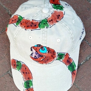 Può includere: Un cappello da baseball beige con un disegno dipinto a mano di un serpente rosso e verde. Il serpente è raffigurato con un occhio blu e una bocca aperta.