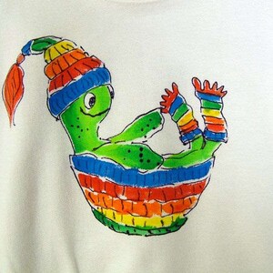May include: White sweatshirt featuring a whimsical, hand-drawn illustration of a green creature with a colorful striped hat and matching striped clothing. The creature is nestled in a striped, rainbow-colored basket. The design is playful and vibrant.