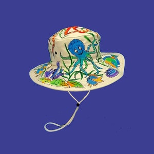 May include: White canvas bucket hat with a hand-painted design of an octopus, fish, and coral. The hat has a black drawstring.