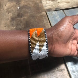 Könnte beinhalten: Ein breites Armband mit Perlen in orangefarbenen, goldenen und weißen geometrischen Mustern. Das Armband hat eine schwarze Lederunterseite und wird am Handgelenk getragen. Ein auffälliges Accessoire.