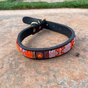 May include: Black leather dog collar with a beaded design in orange, pink, and purple. The collar has a brass buckle and a D-ring for leash attachment. The beaded section is approximately 1 inch wide.