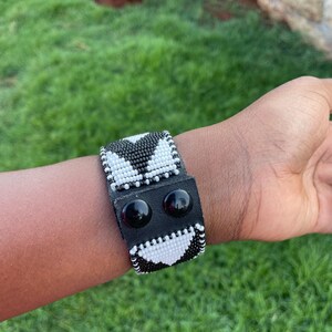 Puede incluir: Pulsera de cuentas en blanco y negro con diseño de corazón. La pulsera tiene una banda de cuero negro con dos cierres de botón a presión negros. La pulsera se lleva en la muñeca.