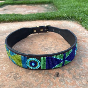 Op de afbeelding: Een zwarte leren hondenhalsband met een kralenpatroon in tinten blauw, groen en turquoise. De halsband heeft een messing gesp en is versierd met geometrische patronen. Het kralengedeelte is ongeveer 3,8 cm breed.