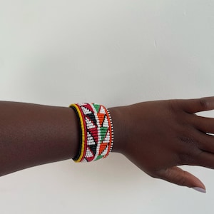 May include: A beaded bracelet featuring a geometric pattern in red, green, white, and orange. The bracelet has a black and yellow border and is worn on a wrist. The design is a vibrant display of color and pattern.