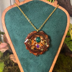 Op de afbeelding: Een ketting met hanger bestaande uit een ronde houten schijf met een glanzende afwerking, ingelegd met een groene ovale edelsteen, een paarse druppelvormige edelsteen, twee gouden maansikkelbedels en twee kleine gouden bloembedels aan een ketting.
