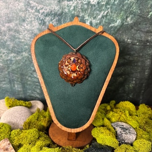 Op de afbeelding: Een bruine dennenappel hanger met een centrale oranje cabochon, een kleine paarse steen erboven en twee goudkleurige maansikkel symbolen aan een donker koord. De hanger wordt gepresenteerd op een houten standaard bekleed met groen fluweel.
