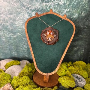 May include: A necklace featuring a circular, dark brown, resin-encased pendant with a polished red stone at the top, a central white stone, and two small silver crescent moon charms on either side, all suspended from a thin, metallic chain.