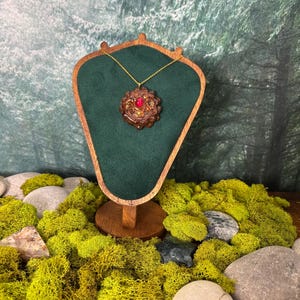 May include: A pendant necklace featuring a brown, textured, circular organic element resembling a pine cone, centered with a smooth, round, vibrant red stone. The pendant hangs from a thin gold-toned chain on a dark green, shield-shaped display.
