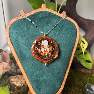 Op de afbeelding: Een ronde hangerketting met een gepolijste dennenappelschaal in hars, ingelegd met drie iriserende maansteen cabochons, kleine zilverkleurige ster- en maansikkelbedels, en een zilverkleurige ketting, gepresenteerd op een houten standaard.