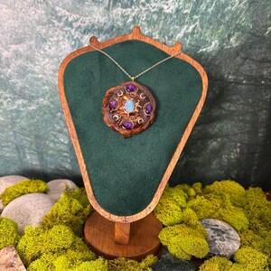 May include: A handcrafted pendant necklace with a circular wooden pendant. The pendant features a central light blue stone, surrounded by purple stones and gold-colored accents. The necklace is displayed on a green velvet-covered stand.